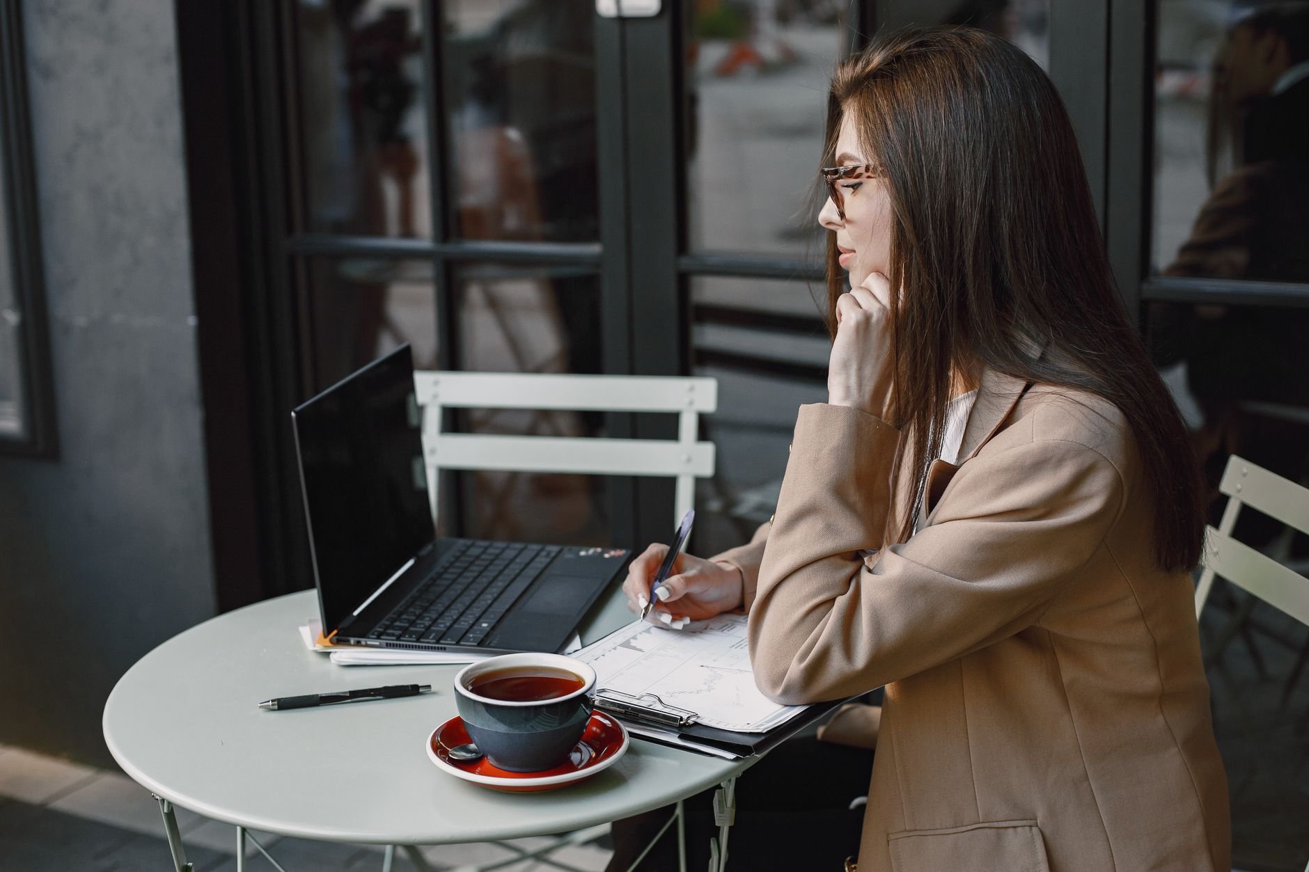 Woman writing in notebook at a coffee shop, using a laptop, with coffee.